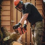 Worker using a chainsaw to cut wood, wearing safety gear in an outdoor setting.